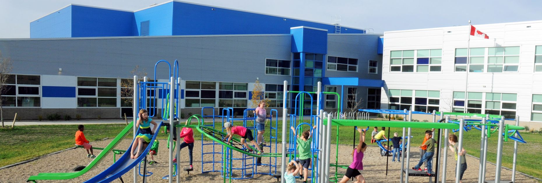 numerous kids in bright t-shirts play on a blue, green and silver playground structure with a modern new school in the bakcground
