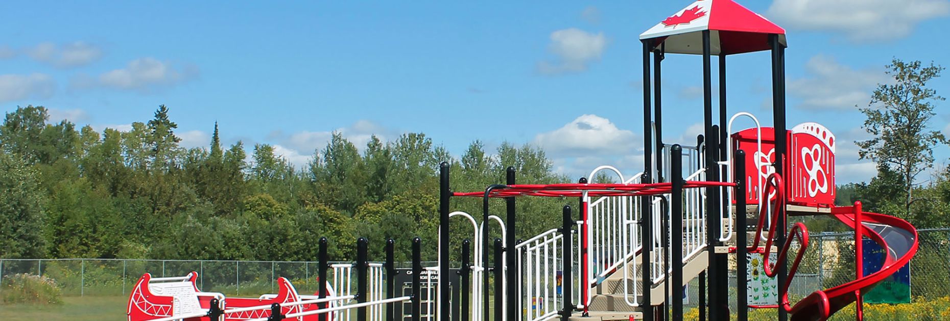 Red and white Blue Imp playground structure with wheelchair ramp leading to the Tippy Canoe, along with overheads, climbers and slides and a hex roof with a maple leaf design. Situated on green turf surfacing with fencing and trees in the background on a blue-sky day.