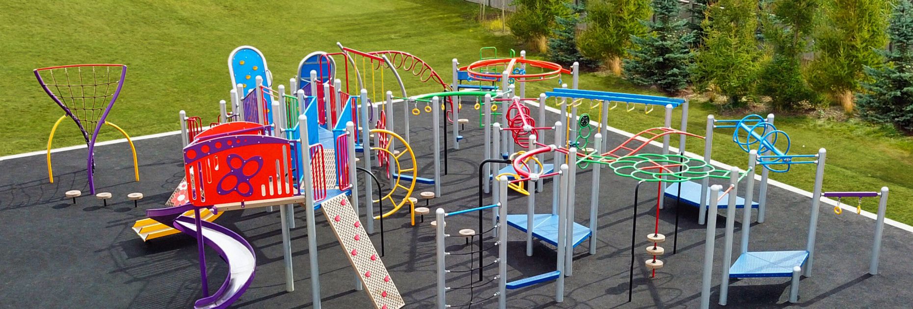 Super colourful, action-packed Blue Imp playground structure with an extensive upper body circuit, diverse climbers and slides. Situated on rubber surfacing with greenery in the background.