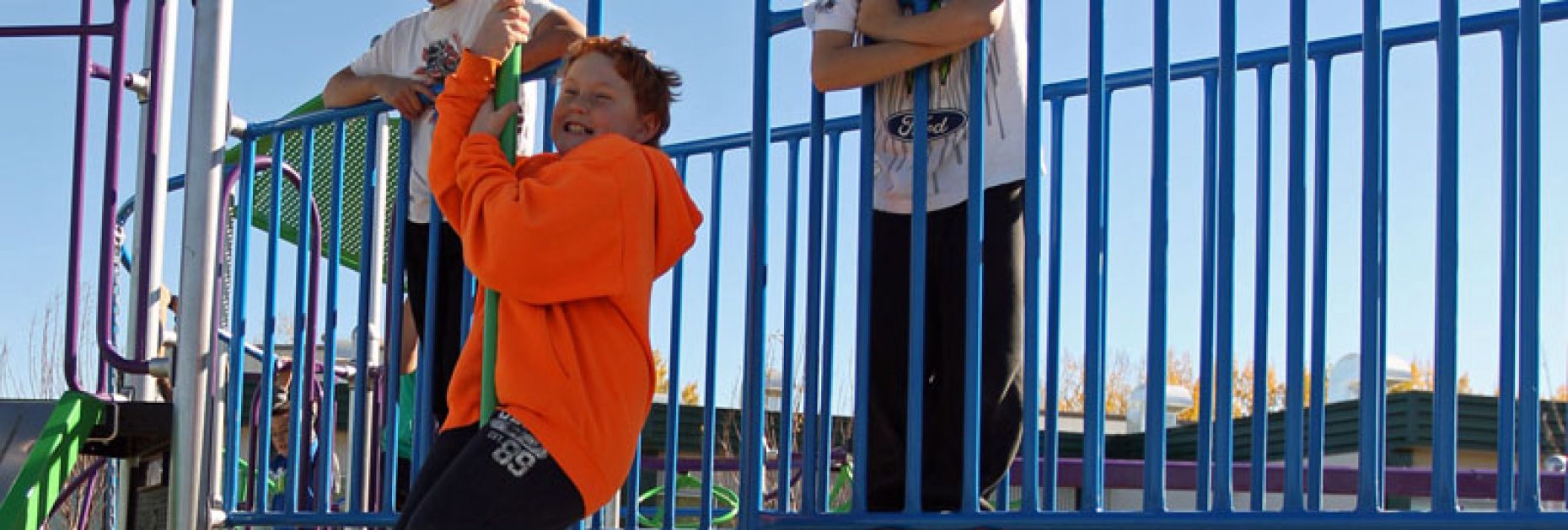 boy in orange hoodie slides down a Fireman's Pole as another boy looks on, on a Catwalk Bridge at a school palyground