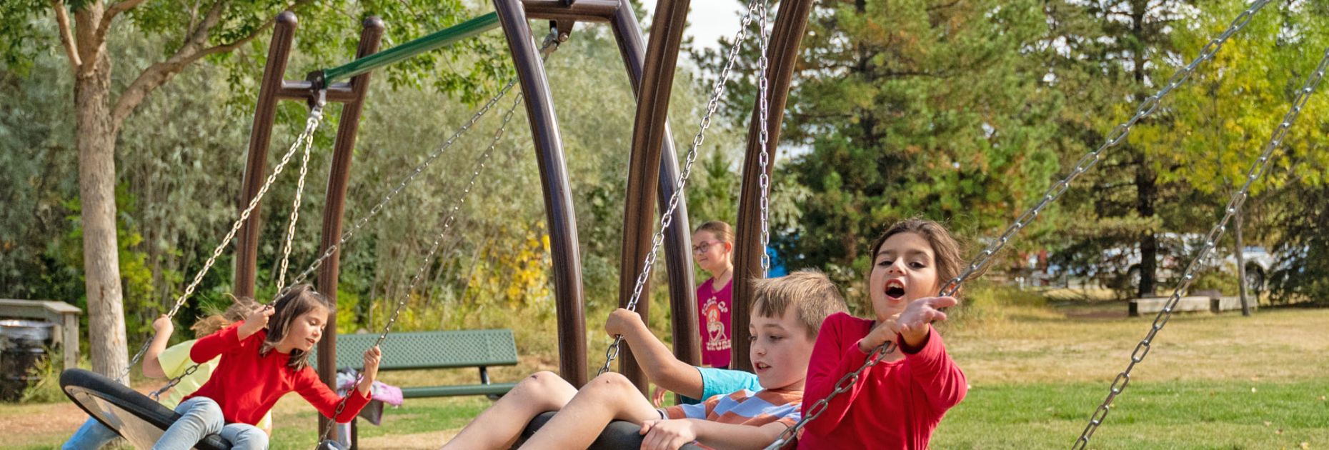two groups of kids swinging on the large, round multi-user swings at a school playground, with trees in the background