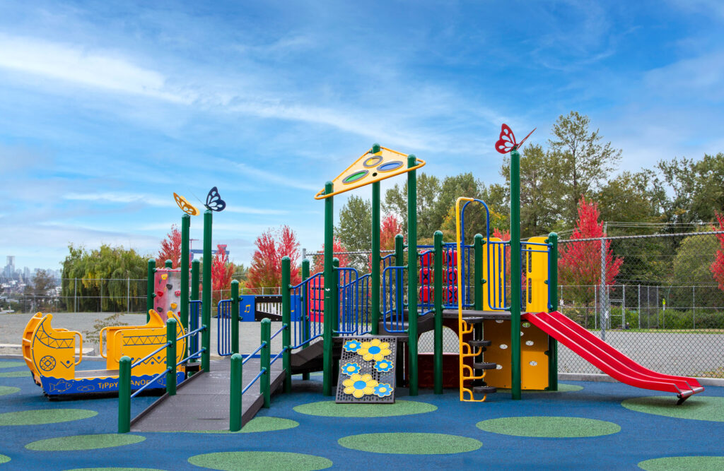 Colourful Blue Imp with rubber surfacing, a wheelchair ramp leading to a yellow Tippy Canoe, various climbers and slides in reds and yellows, topped with bright butterfly post-toppers and a triangle-shaped roof with fall foliage and blue sky in the background at Westview Elementary School in North Vancouver, B.C.